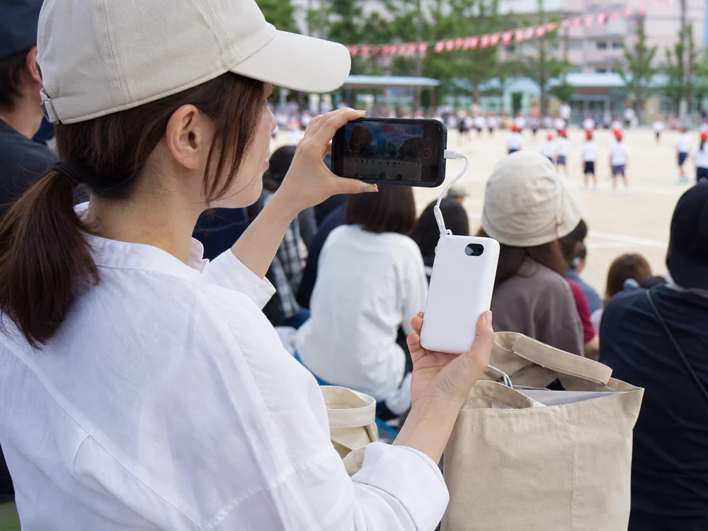 運動会の観覧席でスマホを使いながら白いモバイルバッテリーで給電する様子