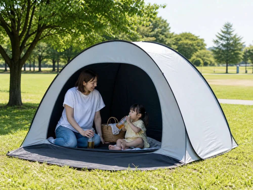 真夏の公園でグレーのポップアップテントの中と前で家族が日差しを避けて休んでいる様子