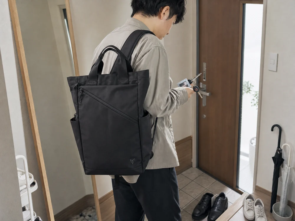 黒いトートリュックを肩にかけた通勤前の人が玄関で持ち物を整えているシーン