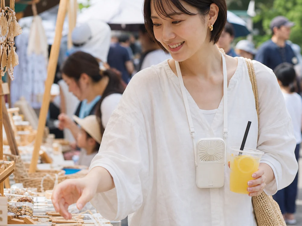 夏の屋外イベントで白いハンディファンを首から下げて使う女性
