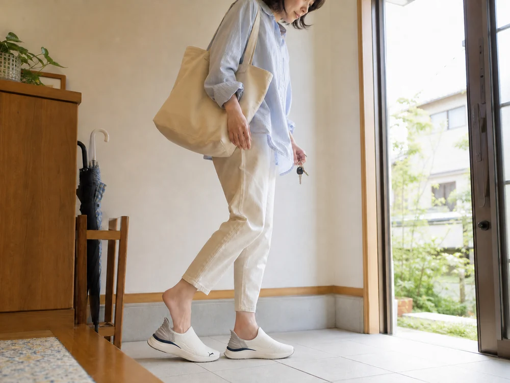 玄関でバッグと鍵を持ちウォームホワイトのスリッポンシューズを履く女性