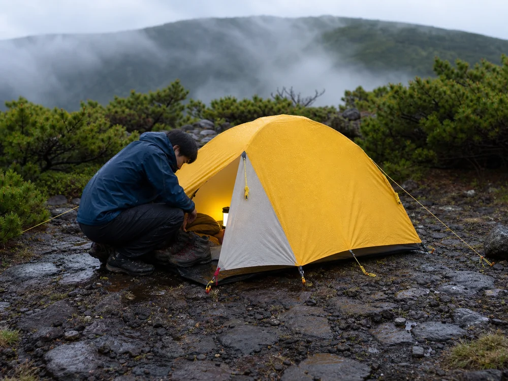 雨上がりの山で黄色いテントの前室に濡れた登山靴を収める登山者