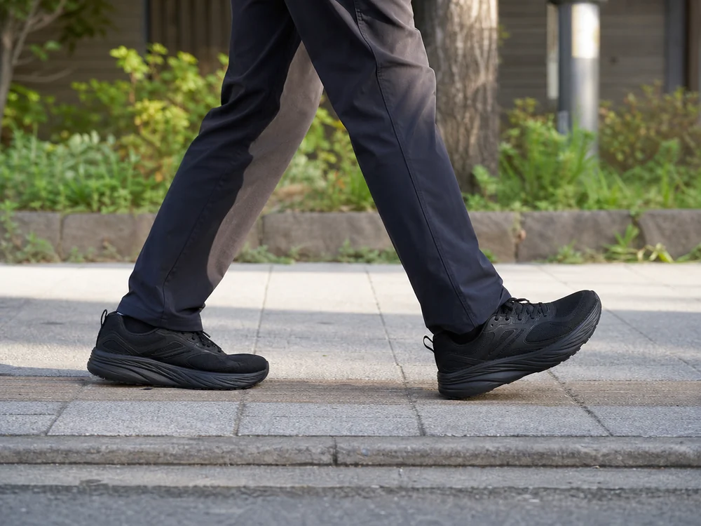 街の歩道をオールブラックの幅広ランニングシューズで歩く男性の足元