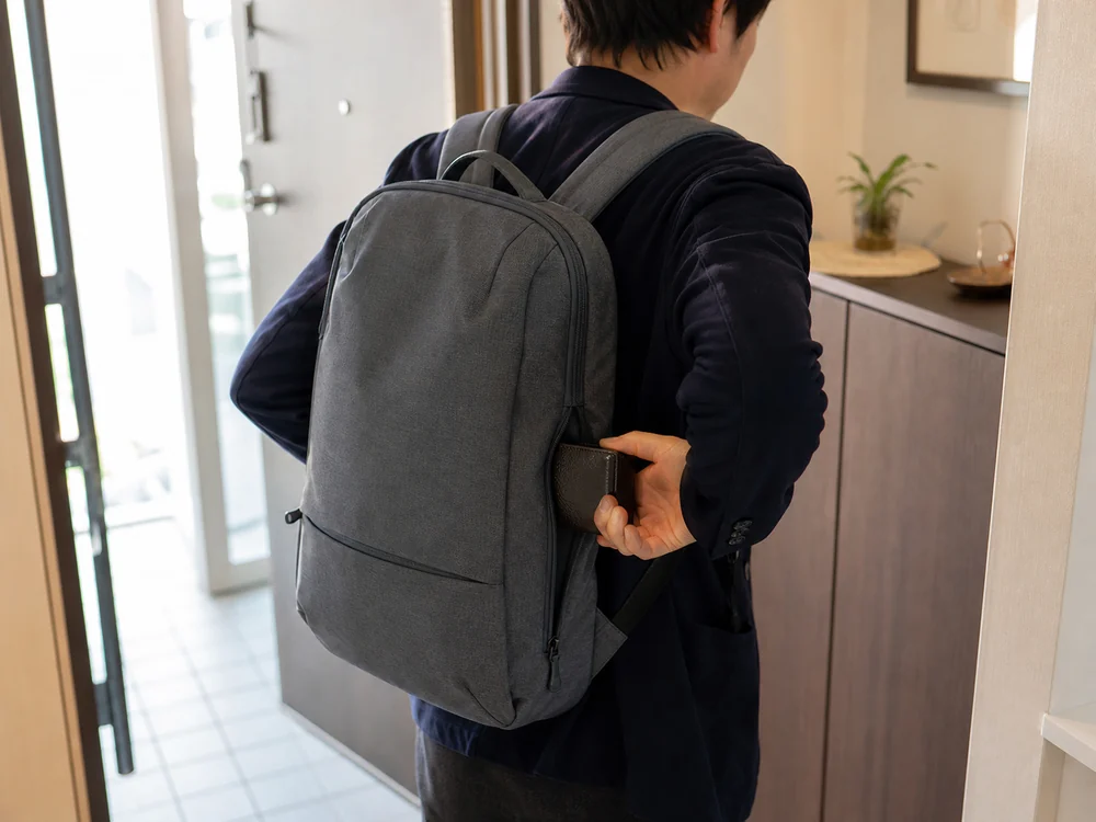 玄関でグレーのバックパックを背負ったまま背面ポケットから財布を取り出す人